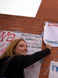 A woman on the CSULB campus puts a banner up on a wall with many posters