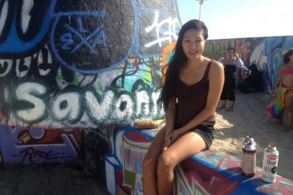 Savannah Cheung sitting on a wall at Venice Beach next to her name "Savannah" painted on the wall in white bubble letters with a black outline