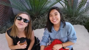 Marina Barnes & Eujin Song sitting on a stone in the CSULB School of Art, Art Gallery Courtyard. Both with smiles. Barnes wearing sunglasses.