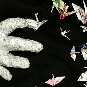paper hand reaching toward many cranes "flying" around against a black background