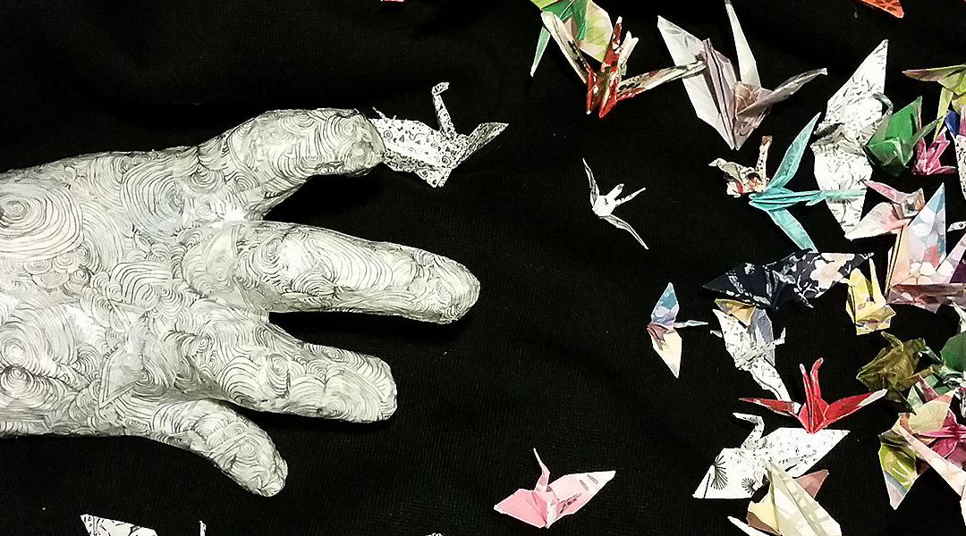 paper hand reaching toward many cranes "flying" around against a black background